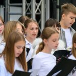 Grupa dzieci śpiewa w chórze na scenie, w białych strojach. Za nimi widać konstrukcję aluminiową sceny. Dzieci skupione na występie.