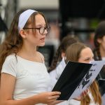 Na zdjęciu dziewczynka w białej koszulce i opasce trzyma kartkę z portretem. W tle widać inne dzieci. Scena jest w jasnych kolorach.