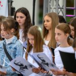 Grupa młodych dziewcząt w białych koszulkach stoi na scenie, trzymając kartki z czarno-białymi zdjęciami. W tle widoczny jest mikrofon i częściowo zarysowany napis. Kompozycja jest jasna i dynamiczna.