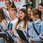 Grupa dzieci śpiewa, trzymając nuty z czarno-białymi portretami. Są w jasnych ubraniach. W tle logo i mikrofon.