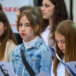 Grupa młodych osób, ubrana głównie na biało, śpiewa na scenie. Środkowa dziewczyna ma jeansową kurtkę i skupiony wyraz twarzy. W tle widać niewyraźne kształty i napis.