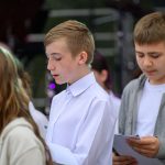 Grupa młodych osób stoi i czyta z kartek. Mają na sobie białe koszule i szare bluzy. W tle niewyraźny sprzęt muzyczny.