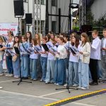 Grupa młodych osób śpiewa na scenie na świeżym powietrzu. Ubrani są w białe bluzki i dżinsy. W tle widoczne są instrumenty muzyczne i baner z napisem Mazowsze.