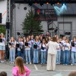 Dzieci śpiewają na scenie na otwartym powietrzu. Mają na sobie białe i niebieskie ubrania, trzymają czarne teczki. W tle są bębny i banery z napisami Powiat Miński i Mazowsze.