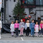 Na scenie dzieci śpiewają w kolorowych strojach. W tle instrumenty muzyczne i baner Mazowsze. Szare, zewnętrzne otoczenie.
