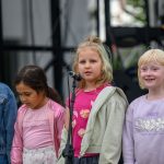 Cztery dziewczynki stoją na scenie przed mikrofonami. Mają różnokolorowe ubrania, jak denimowa kurtka i różowe bluzy. Tło to metalowa konstrukcja z delikatnym zarysem roślinności.