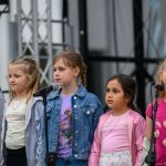 Na scenie pięć dziewczynek śpiewa przy mikrofonach. Mają kolorowe ubrania, m.in. różowe i niebieskie. W tle widać konstrukcję sceny.
