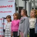 Pięć dziewczynek śpiewa na scenie na tle baneru z napisem Mazowsze. Ubrane są kolorowo, stoją przy mikrofonach. Tło jest jasne.