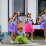 Grupa dziewczynek w kolorowych spódniczkach tańczy na zewnątrz. Tło to biały budynek z oknami. Ich stroje są jaskrawe i radosne.