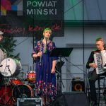 Trzy osoby grają muzykę na scenie. Kobieta w ludowej sukience gra na klarnecie, obok mężczyźni z bębnem i akordeonem. Tło z bębnem i zielenią.
