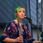 Kobieta w niebieskiej sukni z kolorowymi kwiatami gra na klarnecie przy mikrofonie. Tło jest rozmyte, ciemno-szare. Włosy blond z zieloną poświatą.