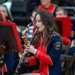 Orkiestra dęta gra na świeżym powietrzu. Muzycy mają czerwono-niebieskie mundury. Na pierwszym planie kobieta gra na klarnecie.