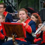 Grupa młodych muzyków w czerwono-granatowych uniformach gra na trąbkach. Tło ciemne, sceniczne, z elementami sprzętu muzycznego. Atmosfera koncertu na świeżym powietrzu.