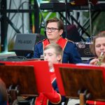 Grupa muzyków w granatowo-czerwonych mundurach na scenie. Mężczyzna z okularami siedzi z poważnym wyrazem twarzy. W tle sprzęt nagłaśniający.