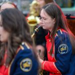 Orkiestra dęta w kolorowych, granatowo-czerwonych mundurach. Muzycy grają na instrumentach dętych. Tło rozmyte.