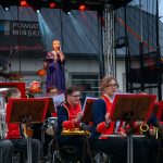 Scena plenerowa z orkiestrą w czerwono-niebieskich mundurach. Kobieta w granatowej sukni przemawia. W tle szyb na budynku.
