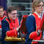 Muzycy w orkiestrze w czerwono-granatowych mundurach grają na instrumentach. Na pierwszym planie saksofon. Tło jest rozmyte.
