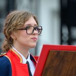 Osoba w okularach i mundurze orkiestry marszowej, z czerwonym szalikiem. W tle rozmyte szczegóły budynku. Kolory to głównie czerwień i granat.