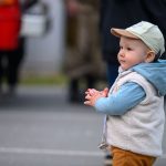 Małe dziecko w czapce i niebieskiej bluzie stoi na zewnątrz. Tło jest rozmyte, widać czerwony kształt. Dziecko ma zawieszkę na smoczku.