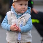 Chłopiec w beżowej czapce, niebieskiej bluzie i białej kamizelce z pluszowym materiałem. Ma brązowe spodnie, w tle rozmyte postaci. Trzyma smoczek na klipsie.