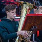 Muzyk gra na tubie podczas koncertu. Instrument mieni się złotym kolorem, a w tle widać sceniczne oświetlenie. Młody mężczyzna nosi zieloną bluzę, a przed nim znajduje się pulpit z nutami.