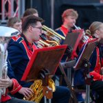 Orkiestra dęta w kolorowych, granatowo-czerwonych uniformach. Muzycy grają na instrumentach, głównie saksofonach. Tło rozmyte, sceniczne oświetlenie.
