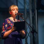 Osoba w granatowej sukience z kwiatowym wzorem przemawia do mikrofonu. Tło to metalowa konstrukcja i ciemne, rozmyte elementy. Światło pada na twarz, podkreślając jej rysy.