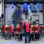 Orkiestra w czerwono-niebieskich mundurach gra na scenie. W tle wiszą reflektory i bębny. Scena ustawiona na zewnątrz.