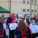 Mężczyzna dyryguje orkiestrą w mundurach z czerwonymi elementami. W tle ludzie siedzą przy stolikach na zewnątrz budynku. Tło jest beżowe z zielonym namiotem.