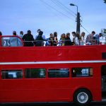 Czerwony piętrowy autobus z otwartym górnym pokładem wypełniony ludźmi. Wieczorne niebo, latarnia i drzewa w tle. W dolnej części kilka osób wsiada lub wysiada.