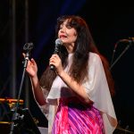 Kobieta śpiewa na scenie w różowo-fioletowej sukni. Trzyma mikrofon, tło jest ciemne, z widocznym sprzętem dźwiękowym. Luminescencja sceny podkreśla jej strój.