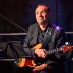 Muzyk w czarnym garniturze siedzi z gitarą basową. Tło jest ciemne, sceniczne, z nutami na stojaku. Wyraz twarzy spokojny.