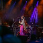 Kobieta w różowej sukience śpiewa na scenie. W tle muzycy i kolory świateł: pomarańczowy i fioletowy. Ciemne tło sceniczne.