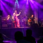 Na scenie oświetlonej fioletowymi światłami występuje wokalistka i gitarzysta. Kobieta ma długą suknię, a mężczyzna siedzi z gitarą. W tle perkusja i pianino, widoczni są także widzowie.