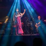 Na scenie kobieta w biało-różowej sukience śpiewa, oświetlona niebieskim i fioletowym światłem. W tle gra gitarzysta. Tło jest ciemne.