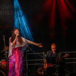 Na zdjęciu piosenkarka w białej bluzce i kolorowej spódnicy śpiewa do mikrofonu. Obok muzyk gra na gitarze basowej. Tło sceny jest ciemne, oświetlone niebieskimi i czerwonymi światłami.