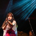 Kobieta w kolorowej sukni śpiewa na scenie, trzymając mikrofon. Za nią perkusista i ciemne tło z jasnymi światłami.