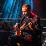 Muzyk gra na gitarze basowej w ciemnym ubraniu. Tło jest niebieskie z jasnym oświetleniem. Obok stojak na nuty.