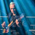 Mężczyzna gra na gitarze elektrycznej na scenie. Oświetlenie w niebieskich odcieniach. Tło ciemne z metalowymi barierkami.