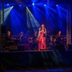 Koncert na scenie, wokalistka na środku w różowej sukni, muzycy po bokach. Oświetlenie niebieskie i fioletowe. Tło ciemne.