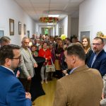 Grupa ludzi w korytarzu, większość w eleganckich strojach. Ściany białe, na nich obrazy. Na suficie kolorowe dekoracje.