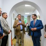 Sześć osób w korytarzu, eleganckie stroje, dyskutują. Ściany jasne, zdjęcia na półkach. Podłoga drewniana, okno w tle.