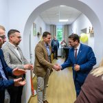 Grupa mężczyzn w garniturach wita się uściskiem dłoni w jasnym korytarzu. Tło to białe ściany z obrazami. Klimat formalny, wydarzenie w pomieszczeniu.