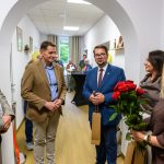 Grupa osób stoi w wąskim korytarzu, na pierwszym planie kobieta trzyma bukiet czerwonych róż. Ściany są białe, na podłodze drewniany panel. W tle widać okno i kilka obrazów na ścianach.