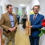 Dwóch mężczyzn stoi w korytarzu. Jeden trzyma torebkę z figurką. Ściany białe, podłoga drewniana, w tle kilka osób.