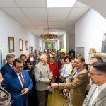 Grupa ludzi stoi w jasnym korytarzu, uśmiechnięci i rozmowni. W tle wiszą kolorowe dekoracje sufitowe. Ściany zdobią obrazy i fotografie.