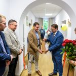 Grupa osób w garniturach i eleganckiej odzieży stoi w jasnym korytarzu. Jeden z mężczyzn przekazuje prezent drugiemu. Po prawej kobieta trzyma bukiet czerwonych róż.