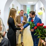 Grupa ludzi w korytarzu wnętrza budynku. Mężczyzna w niebieskim garniturze wręcza czerwone róże kobiecie. W tle jasne ściany i obrazy.