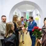 Grupa osób w korytarzu z jasnymi ścianami, dwoje wręcza czerwone róże. Mężczyzna w niebieskim garniturze. Tło z obrazami i żółtym balonem.