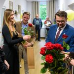 Grupa uśmiechniętych osób wymienia czerwone róże w jasnym pomieszczeniu. Tło z białymi ścianami i jasnymi zasłonami. Obecne elementy dekoracyjne i balon.